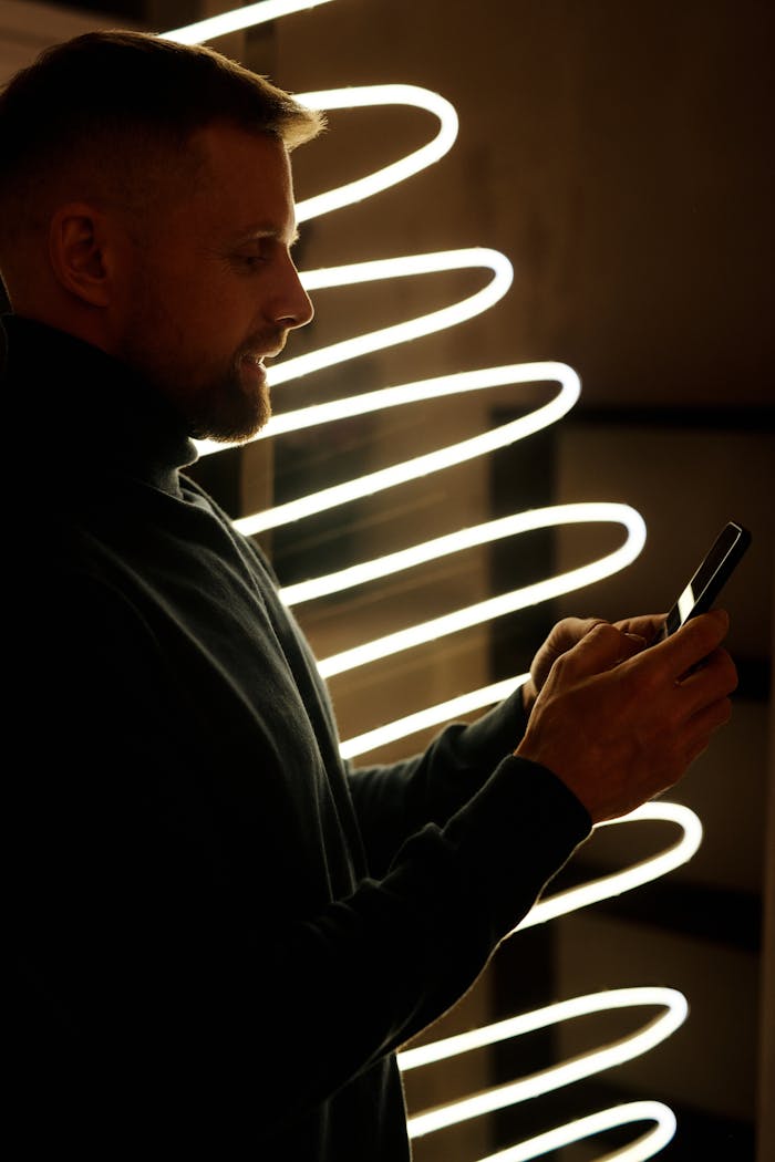 Services A man texting on his smartphone beside festive neon light decor indoors.
